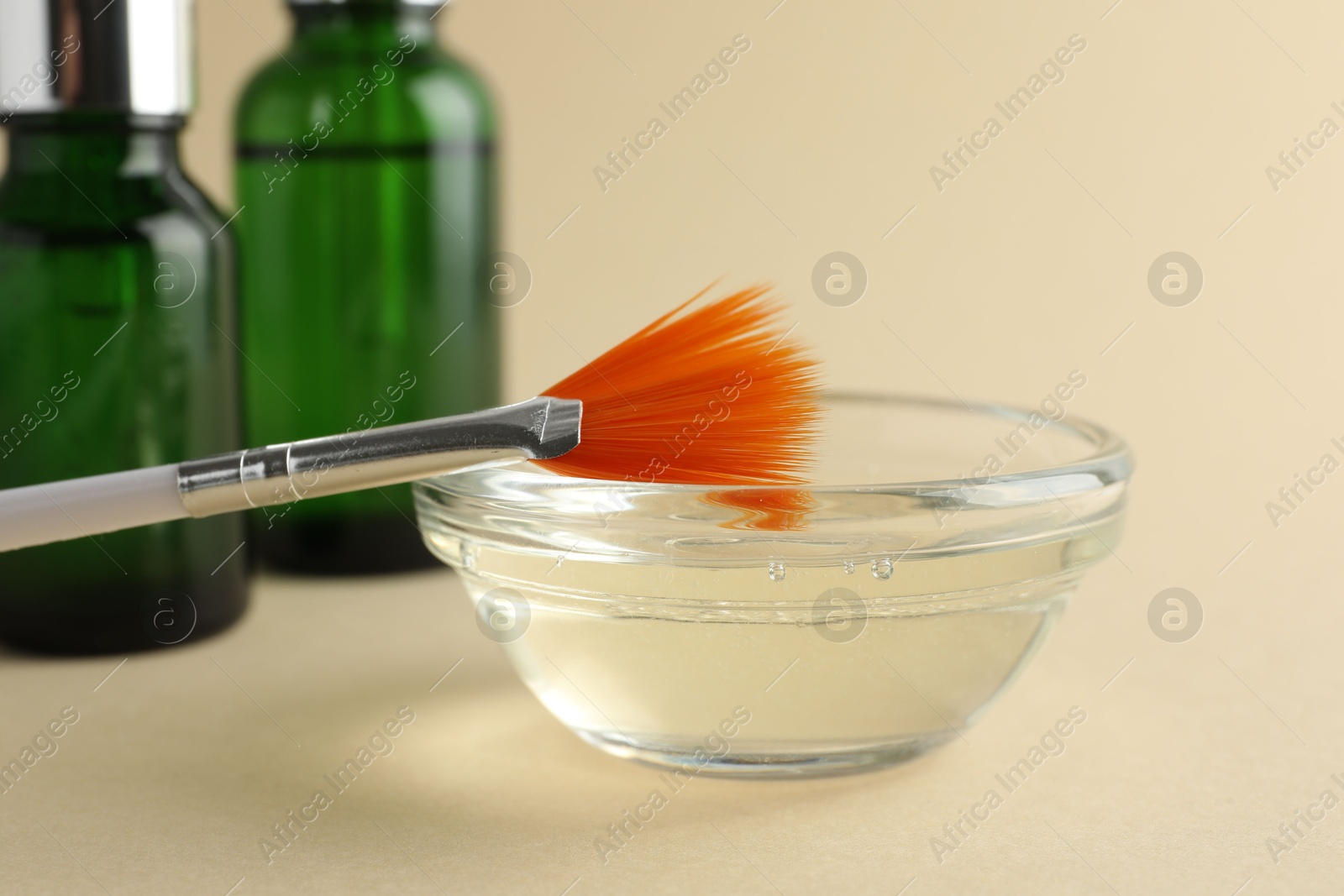 Photo of Chemical peel liquid in bowl, brush and cosmetic bottles on beige background, closeup. Peeling procedure