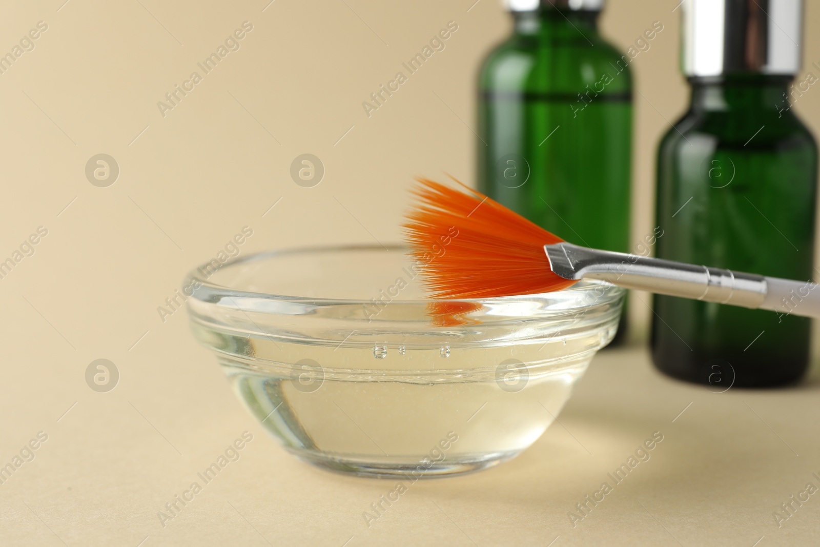 Photo of Chemical peel liquid in bowl, brush and cosmetic bottles on beige background, closeup. Peeling procedure