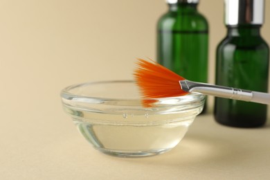 Chemical peel liquid in bowl, brush and cosmetic bottles on beige background, closeup. Peeling procedure