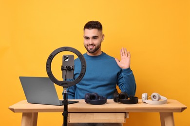 Photo of Technology blogger reviewing headphones and recording video with smartphone and ring lamp at wooden table on orange background