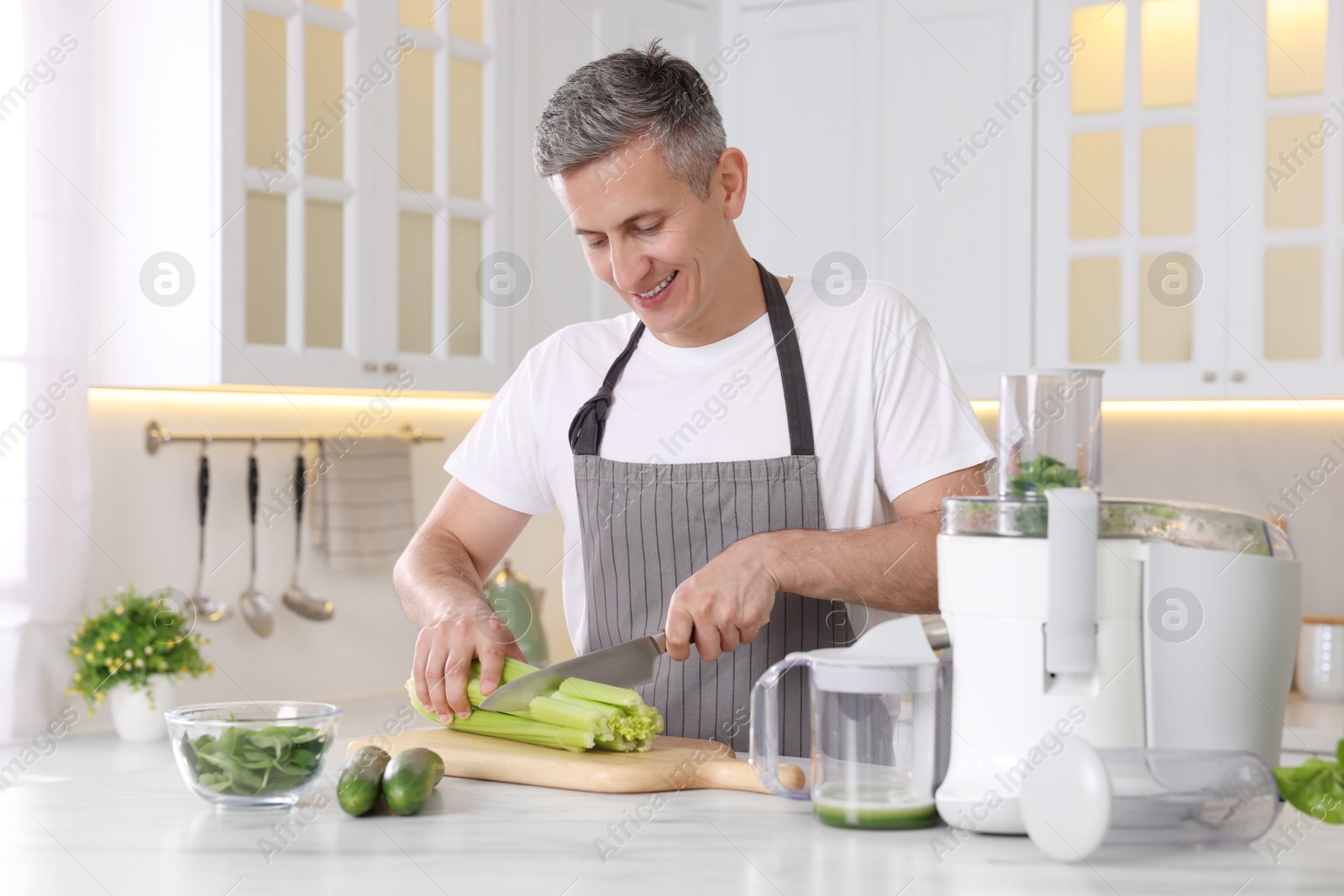 Photo of Juicer and fresh products on white marble table. Smiling man cutting celery in kitchen