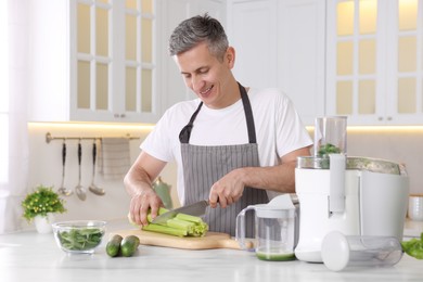 Juicer and fresh products on white marble table. Smiling man cutting celery in kitchen