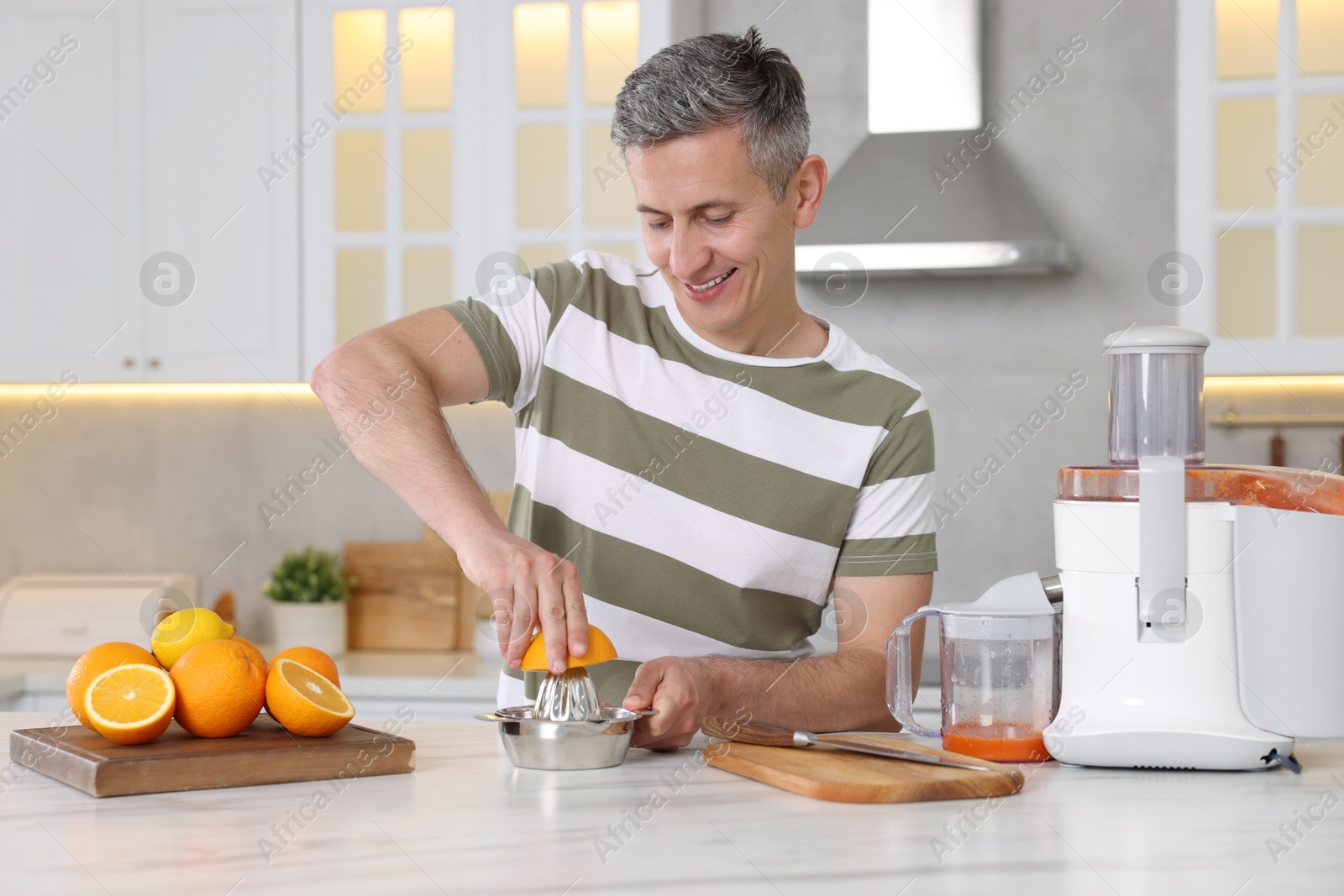 Photo of Smiling man squeezing fresh orange with juicer at white marble table in kitchen