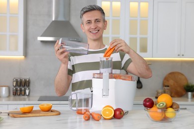 Smiling man putting fresh carrot into juicer at white marble table in kitchen