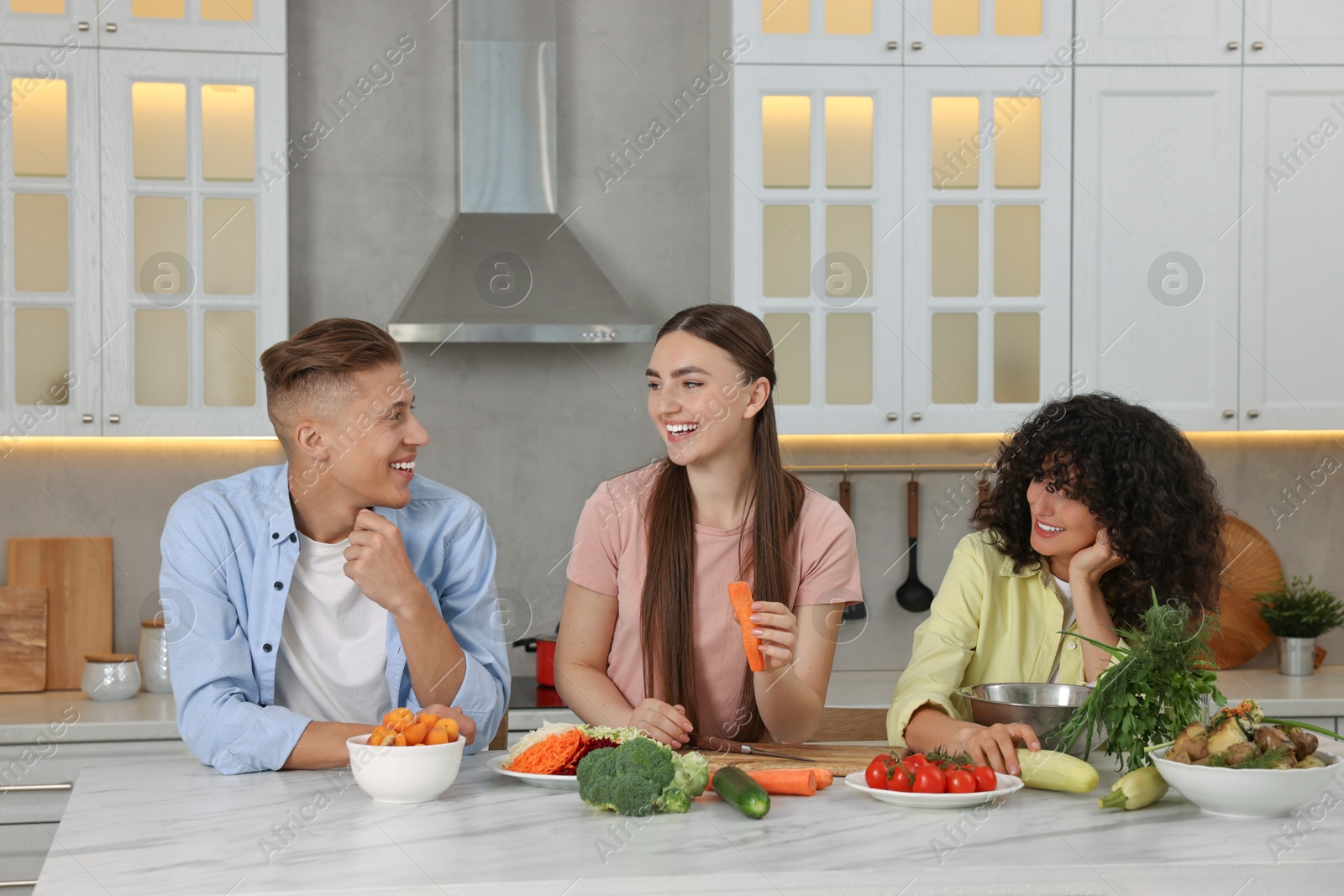 Photo of Friends cooking healthy vegetarian meal at white marble table in kitchen