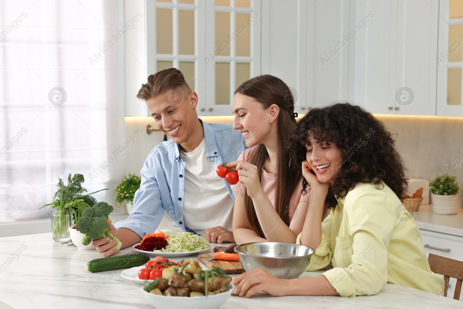 Photo of Friends cooking healthy vegetarian meal at white marble table in kitchen