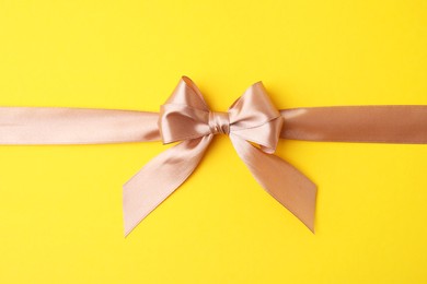 Beige satin ribbon with bow on yellow background, top view