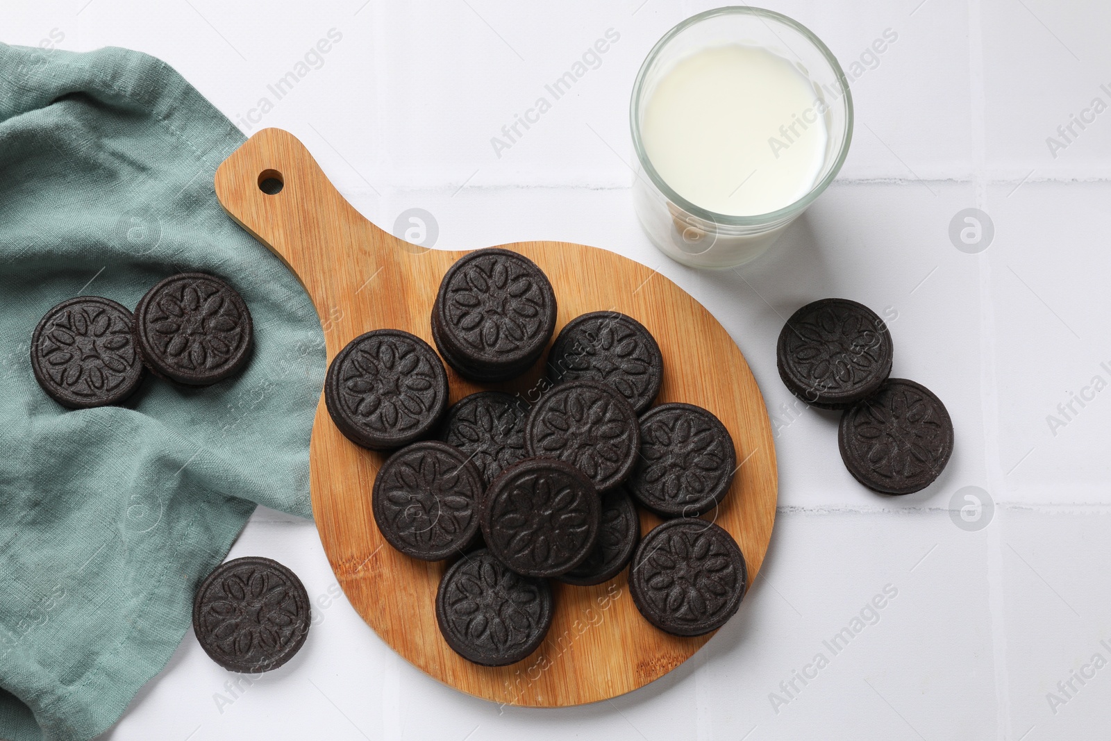 Photo of Board with tasty sandwich cookies and milk on white tiled table, flat lay