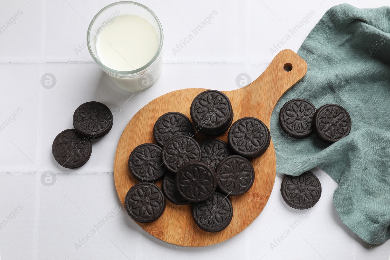 Photo of Board with tasty sandwich cookies and milk on white tiled table, flat lay