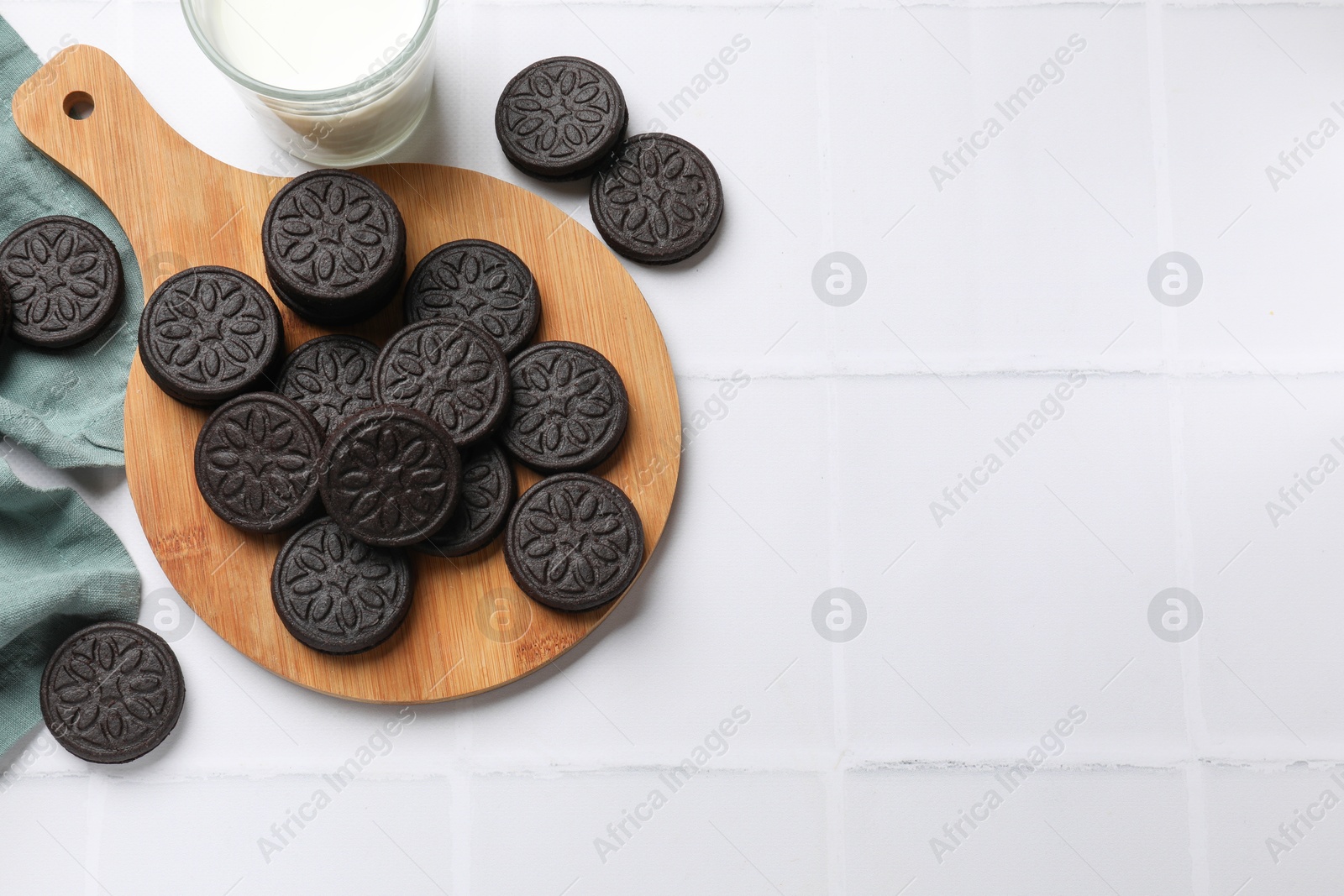 Photo of Board with tasty sandwich cookies and milk on white tiled table, flat lay. Space for text