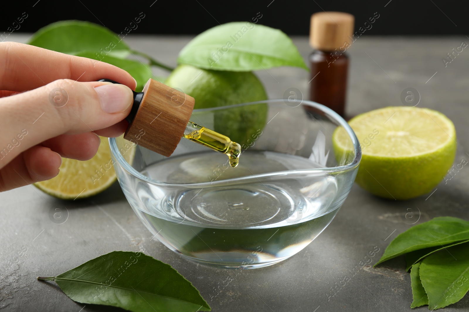 Photo of Woman dripping essential oil from pipette into bowl at gray table, closeup