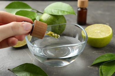 Photo of Woman dripping essential oil from pipette into bowl at gray table, closeup