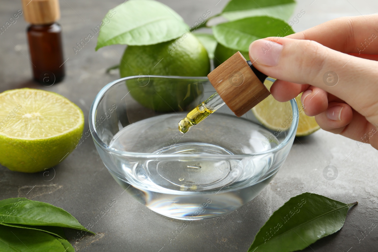 Photo of Woman dripping essential oil from pipette into bowl at gray table, closeup