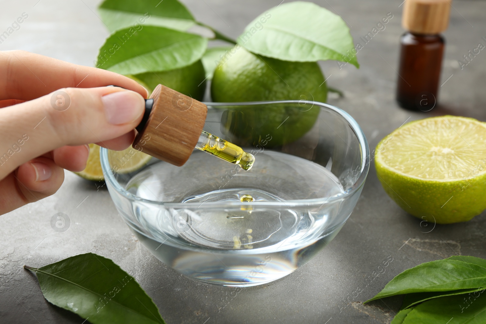 Photo of Woman dripping essential oil from pipette into bowl at gray table, closeup