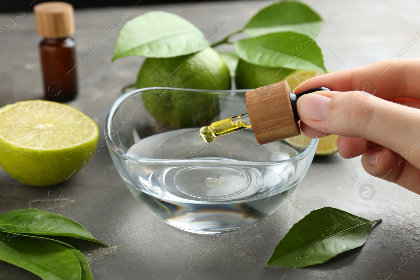 Photo of Woman dripping essential oil from pipette into bowl at gray table, closeup