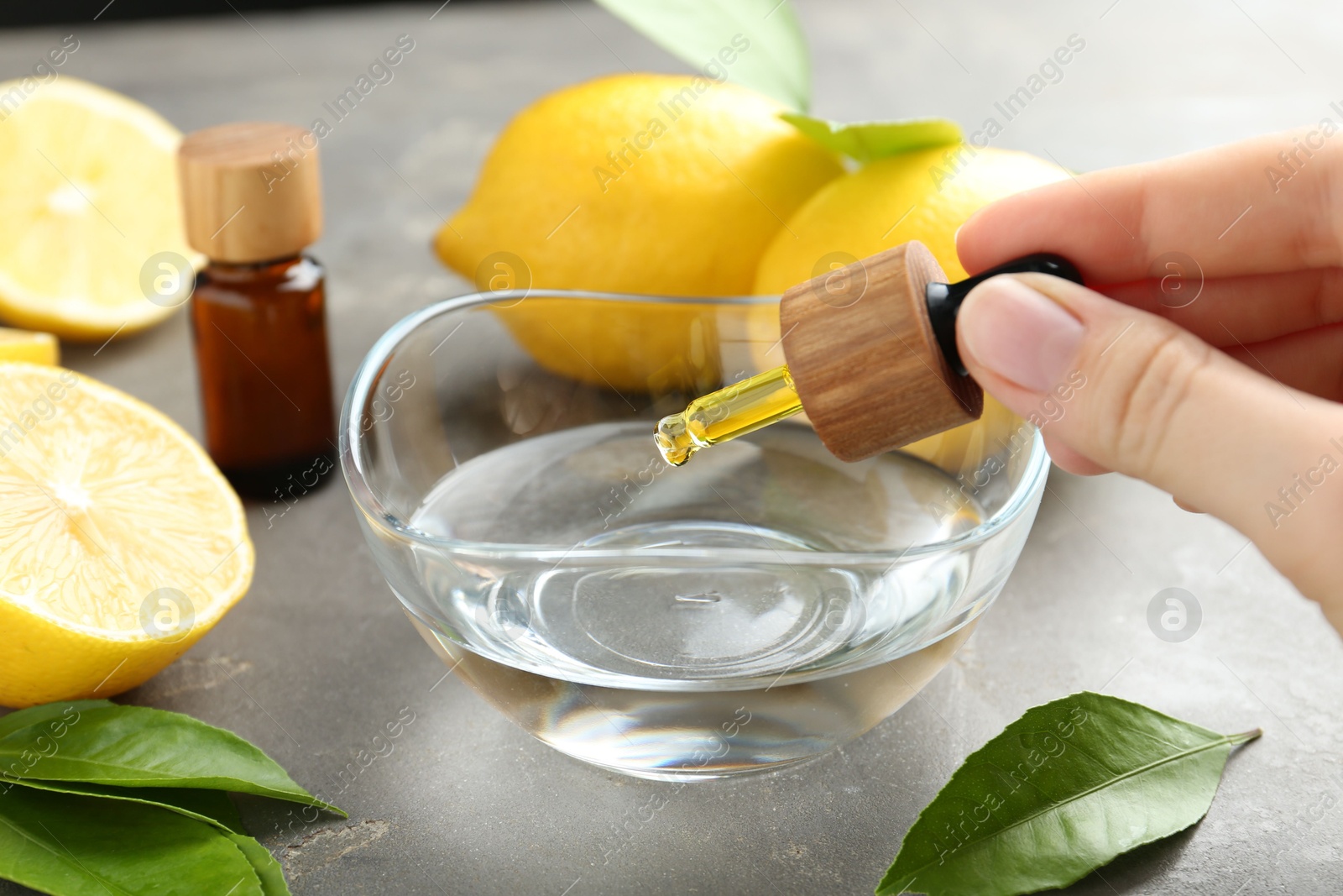 Photo of Woman dripping essential oil from pipette into bowl at gray table, closeup