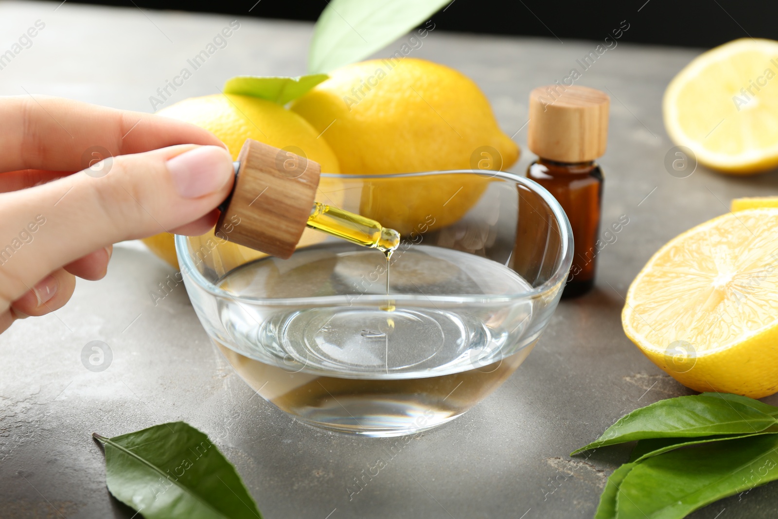 Photo of Woman dripping essential oil from pipette into bowl at gray table, closeup