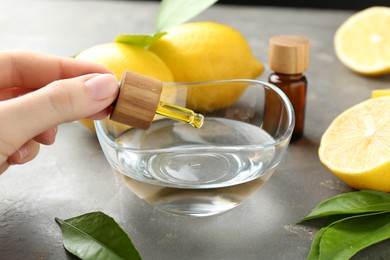 Photo of Woman dripping essential oil from pipette into bowl at gray table, closeup