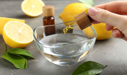 Photo of Woman dripping essential oil from pipette into bowl at gray table, closeup