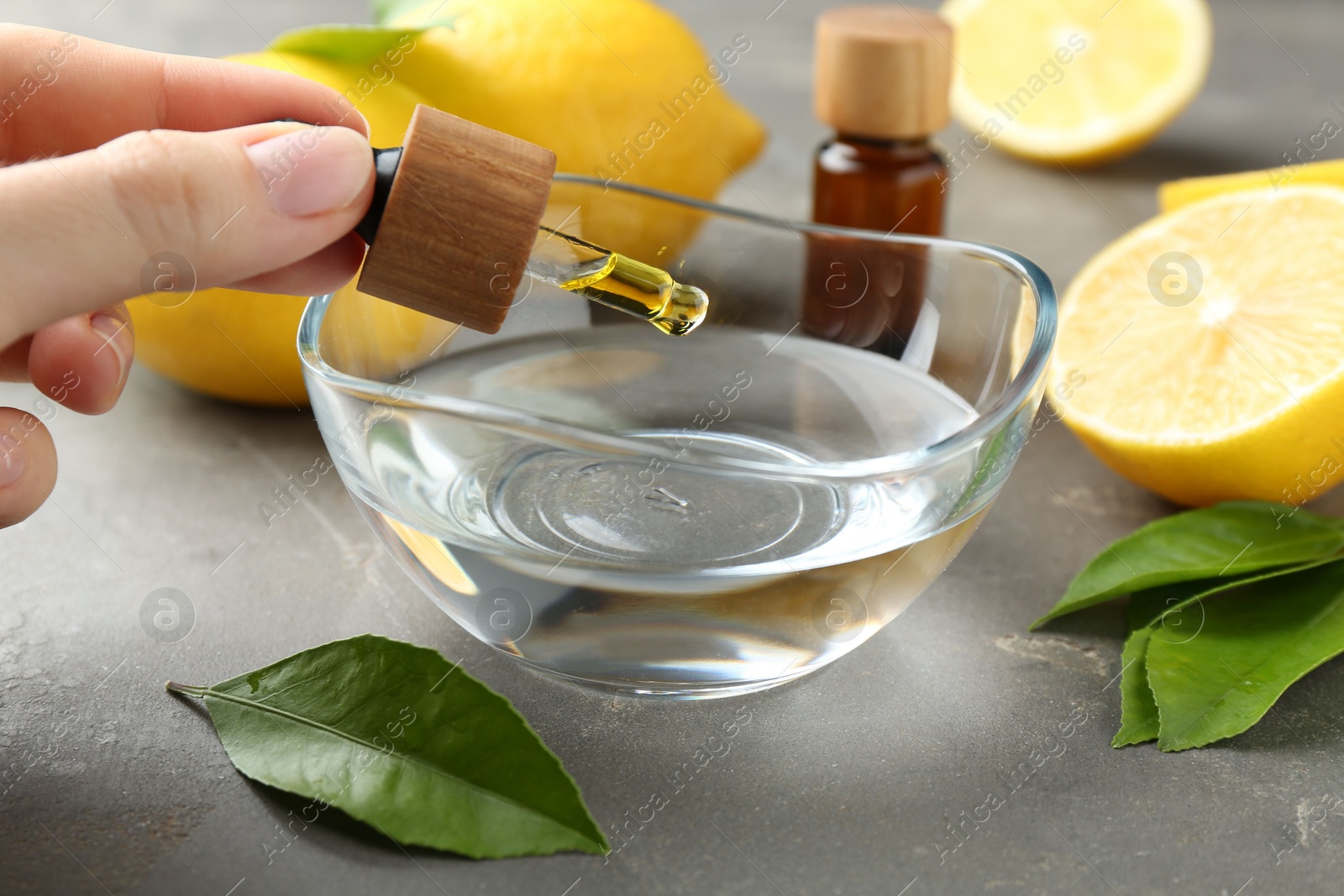 Photo of Woman dripping essential oil from pipette into bowl at gray table, closeup