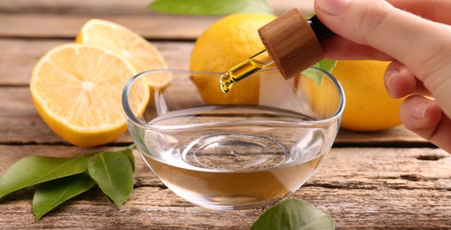 Photo of Woman dripping essential oil from pipette into bowl at wooden table, closeup