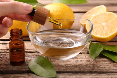 Photo of Woman dripping essential oil from pipette into bowl at wooden table, closeup