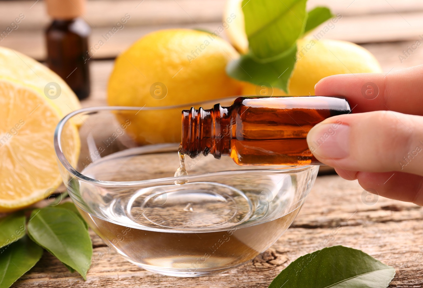 Photo of Woman pouring essential oil from cosmetic bottle into bowl at wooden table, closeup