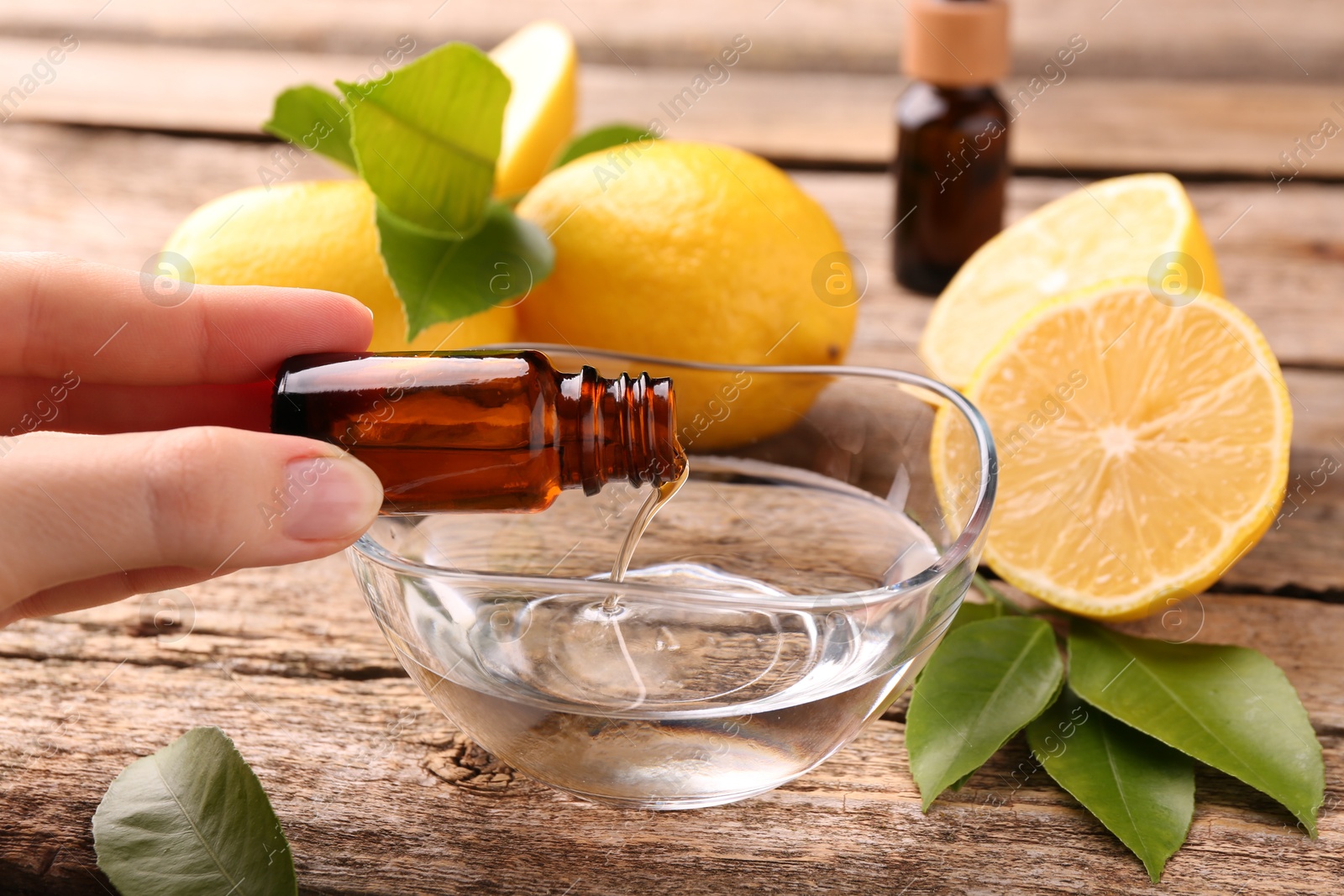 Photo of Woman pouring essential oil from cosmetic bottle into bowl at wooden table, closeup