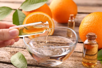 Woman pouring essential oil from cosmetic bottle into bowl at wooden table, closeup