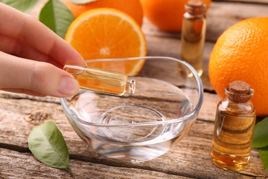 Photo of Woman pouring essential oil from cosmetic bottle into bowl at wooden table, closeup