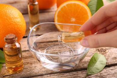 Woman pouring essential oil from cosmetic bottle into bowl at wooden table, closeup