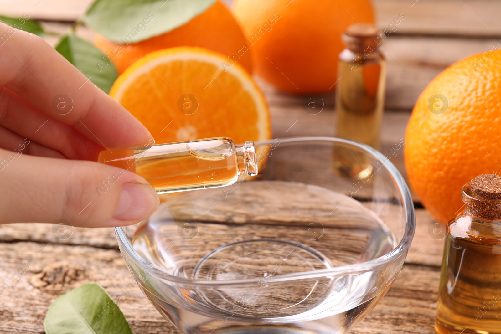 Photo of Woman pouring essential oil from cosmetic bottle into bowl at wooden table, closeup