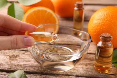 Photo of Woman pouring essential oil from cosmetic bottle into bowl at wooden table, closeup