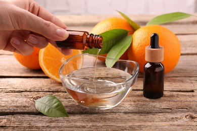 Photo of Woman pouring essential oil from cosmetic bottle into bowl at wooden table, closeup