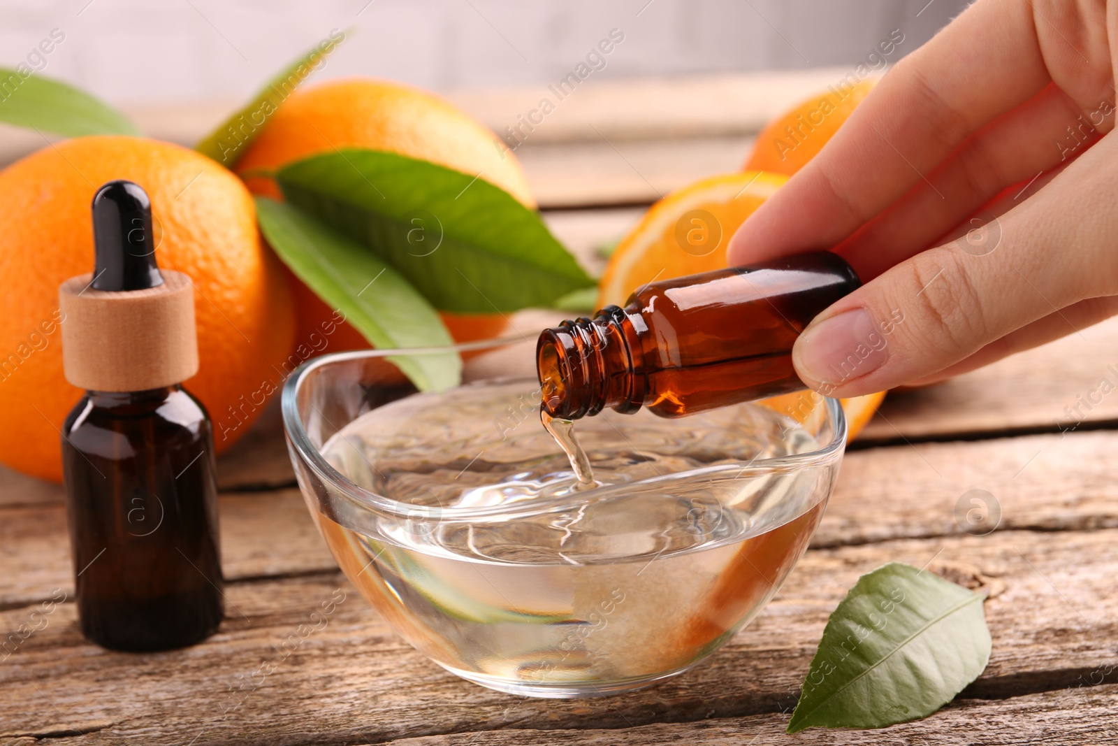 Photo of Woman pouring essential oil from cosmetic bottle into bowl at wooden table, closeup