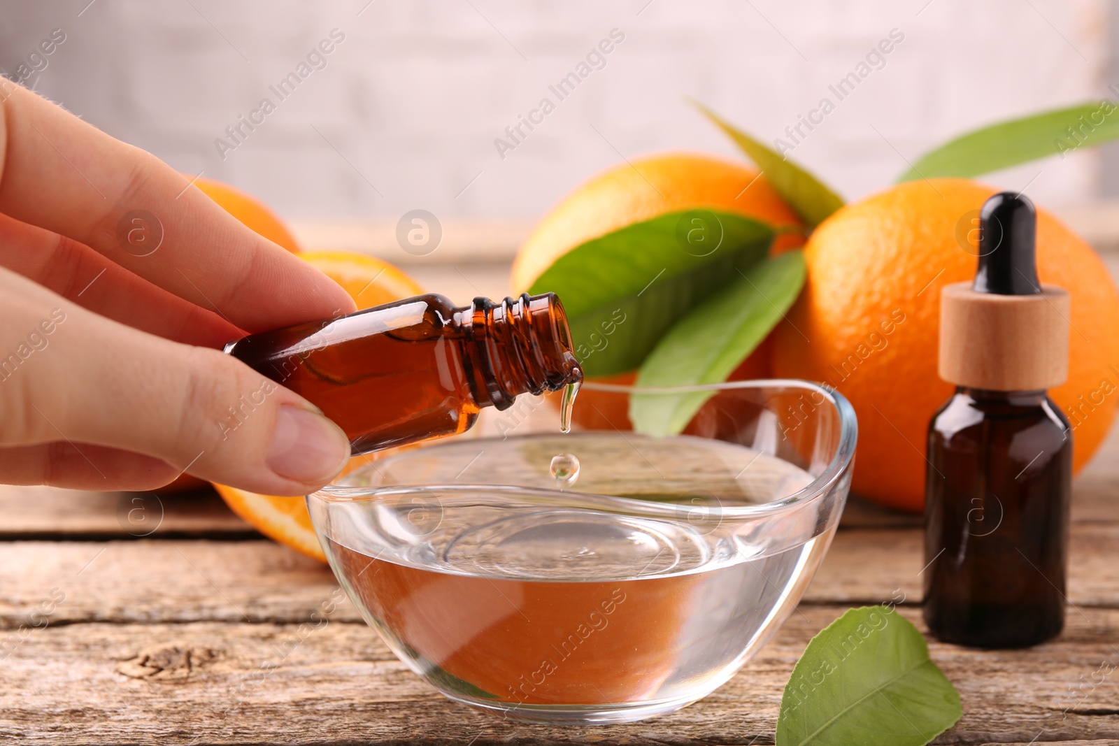 Photo of Woman pouring essential oil from cosmetic bottle into bowl at wooden table, closeup