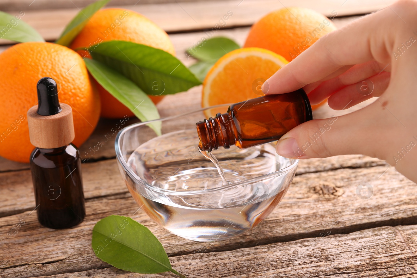 Photo of Woman pouring essential oil from cosmetic bottle into bowl at wooden table, closeup