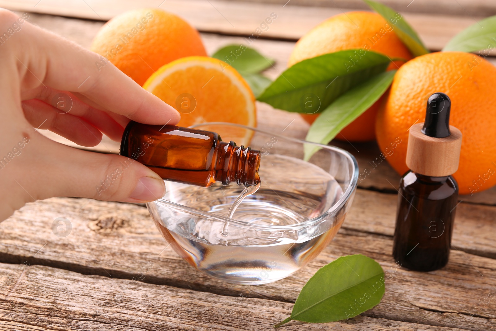 Photo of Woman pouring essential oil from cosmetic bottle into bowl at wooden table, closeup