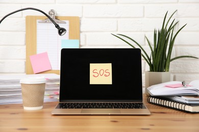 Photo of Note with word SOS, laptop and stationery on wooden table at workplace