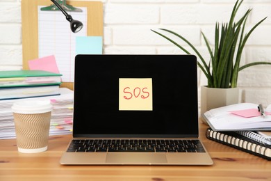 Photo of Note with word SOS, laptop and stationery on wooden table at workplace