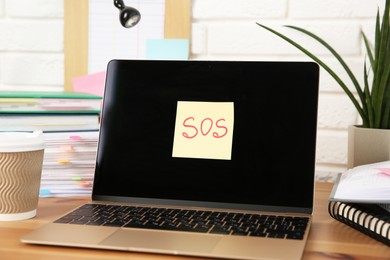 Photo of Note with word SOS, laptop and stationery on wooden table at workplace