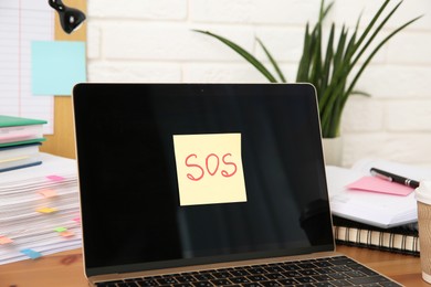 Photo of Note with word SOS, laptop and stationery on wooden table at workplace