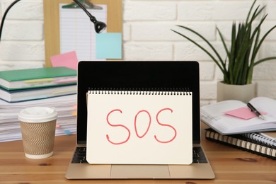 Photo of Notebook with word SOS, laptop and stationery on wooden table at workplace