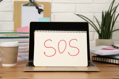 Photo of Notebook with word SOS, laptop and stationery on wooden table at workplace