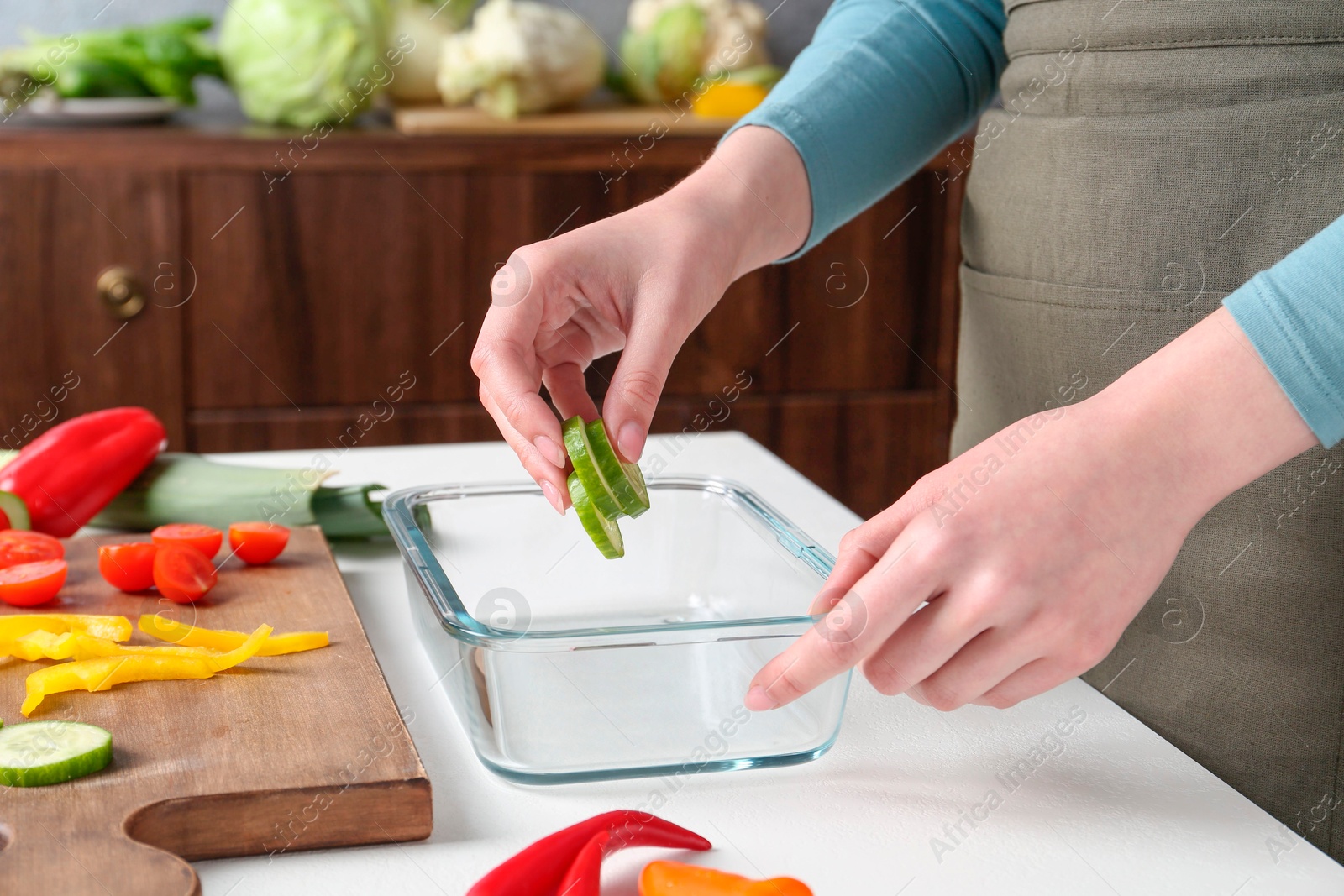 Photo of Healthy food. Woman putting slices of cucumber into glass container at white table in kitchen, closeup