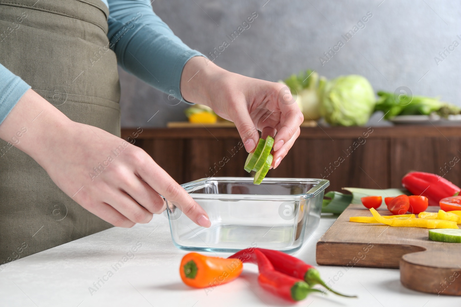 Photo of Healthy food. Woman putting slices of cucumber into glass container at white table in kitchen, closeup