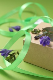 Photo of Beautiful ribbon with flowers and gift box on green background, closeup