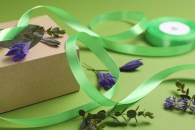Photo of Beautiful ribbon with flowers and gift box on green background, closeup