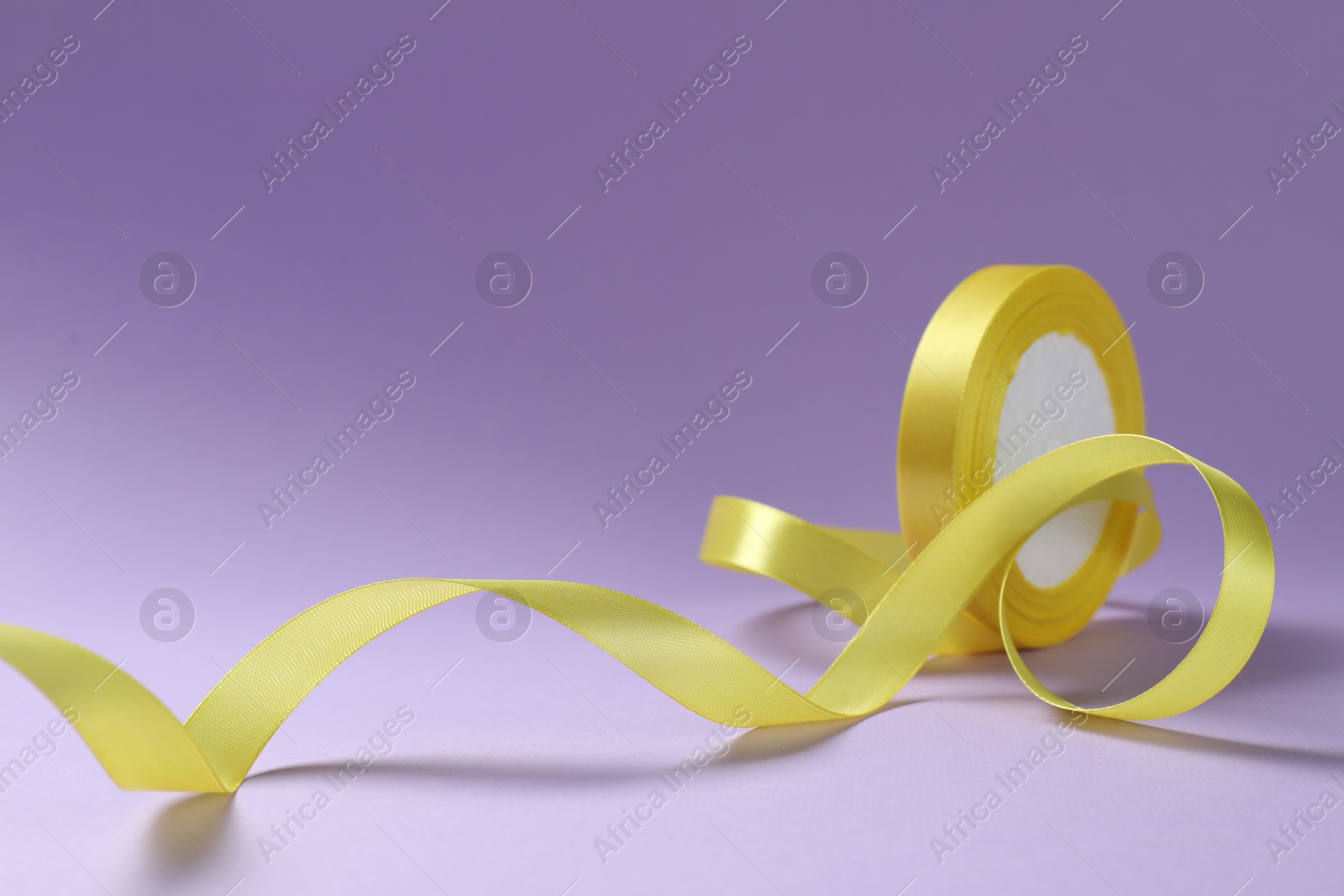 Photo of Beautiful yellow ribbon reel on violet background, closeup. Space for text