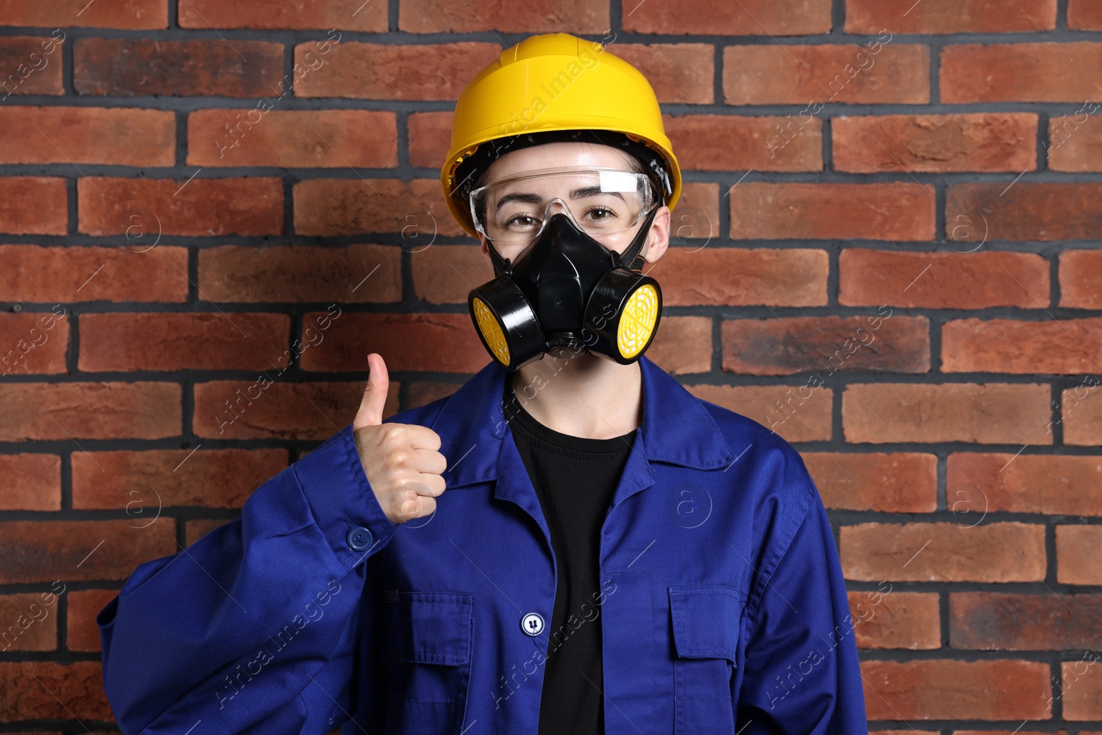 Photo of Worker in respirator, protective glasses and helmet showing thumbs up near brick wall